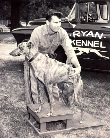 Man posing with a Greyhound at kennel truck.
