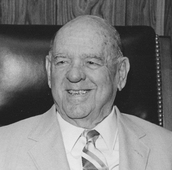collins-jery Smiling man seated in chair, wearing a suit.