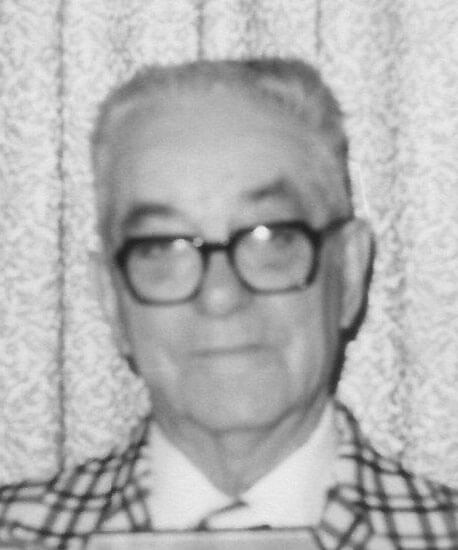 Elderly man wearing glasses, smiling faintly at camera.