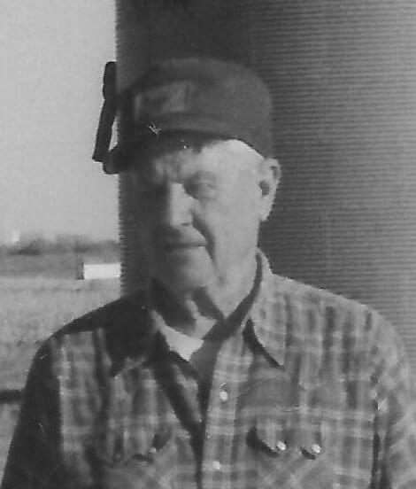 Elderly man in plaid shirt outdoors, wearing cap.