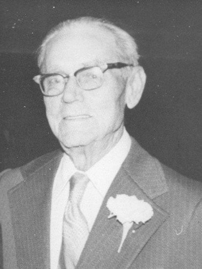 Elderly man wearing glasses, suit, and floral boutonniere.