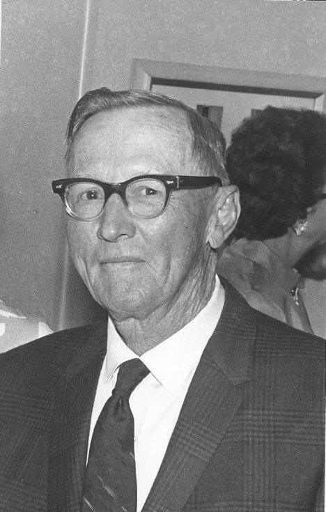 Elderly man in glasses, wearing a suit, smiling.