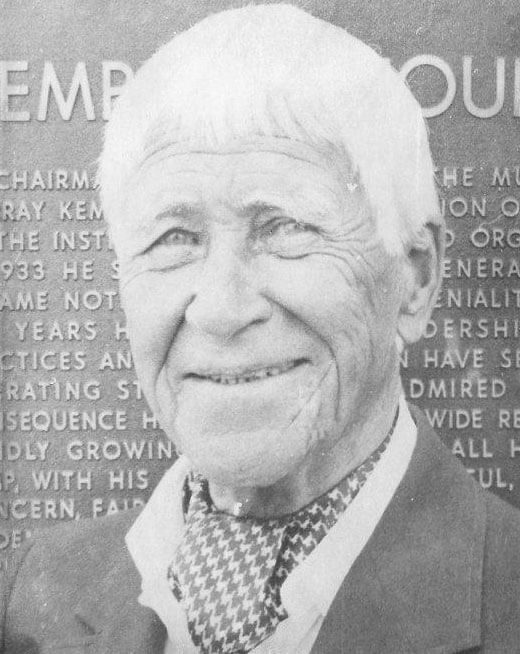 Smiling elderly person in front of engraved plaque.