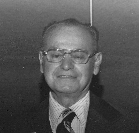Elderly man smiling with glasses and suit.