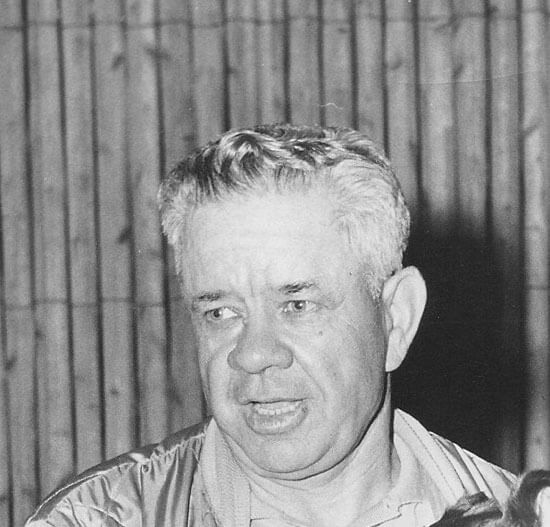 Elderly man speaking in front of wooden backdrop.