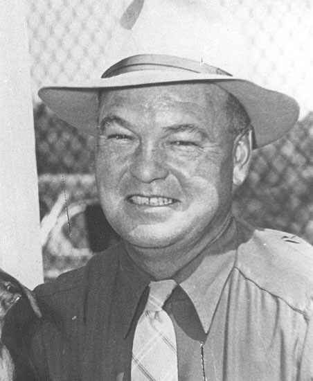 Smiling man in hat poses outdoors by fence.