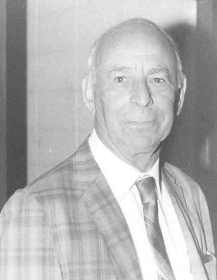 Man in plaid suit, looking forward, smiling slightly.