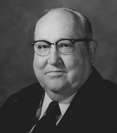 Man in suit and glasses, smiling slightly, black-and-white.