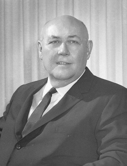 Man in suit seated, looking confidently, with curtains backdrop.
