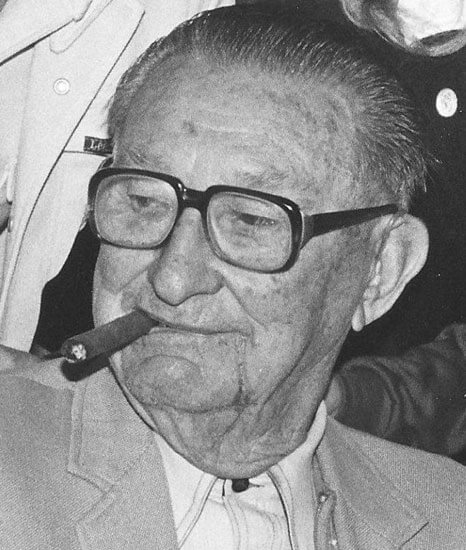 Elderly man with glasses, holding a cigar.